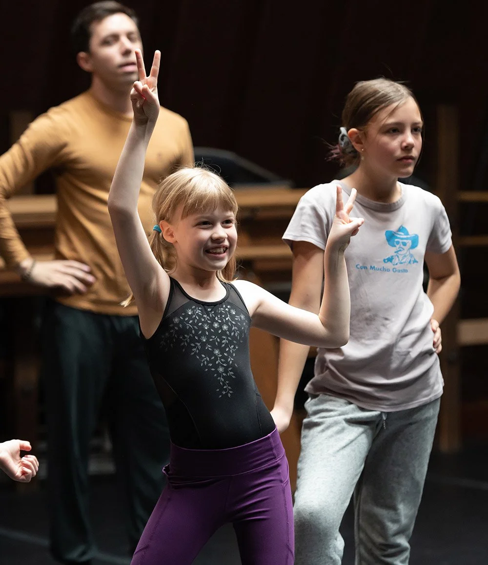 A young girl with blonde hair in a black leotard and purple pants is smiling and making peace signs with both hands. Two other girls and an adult man are in the background, with the older girl in gray and the man in a gold shirt, all in a room with d
