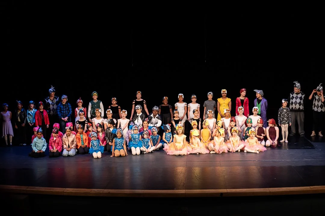 Group of children and adults on stage, dressed in colorful costumes, after a dance performance in a theater.