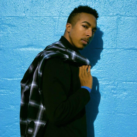 A young man with short hair and a mustache stands against a blue wall, wearing a black shirt and a plaid shirt draped over his shoulder.