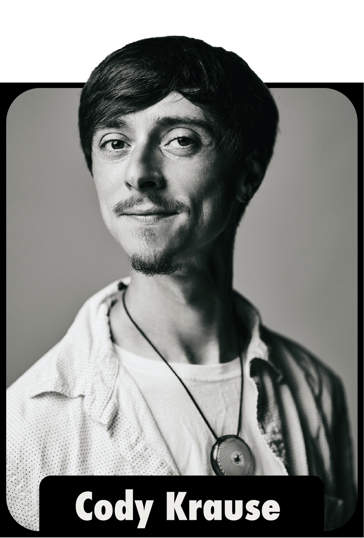 Black and white portrait of Cody Krause with short hair, a goatee, wearing a necklace and a patterned shirt, looking slightly to the right.