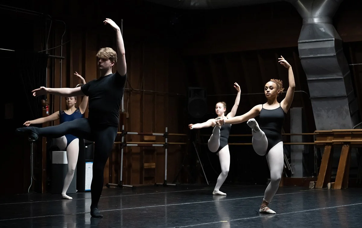Four ballet dancers practicing in a dance studio, wearing black leotards and white tights.