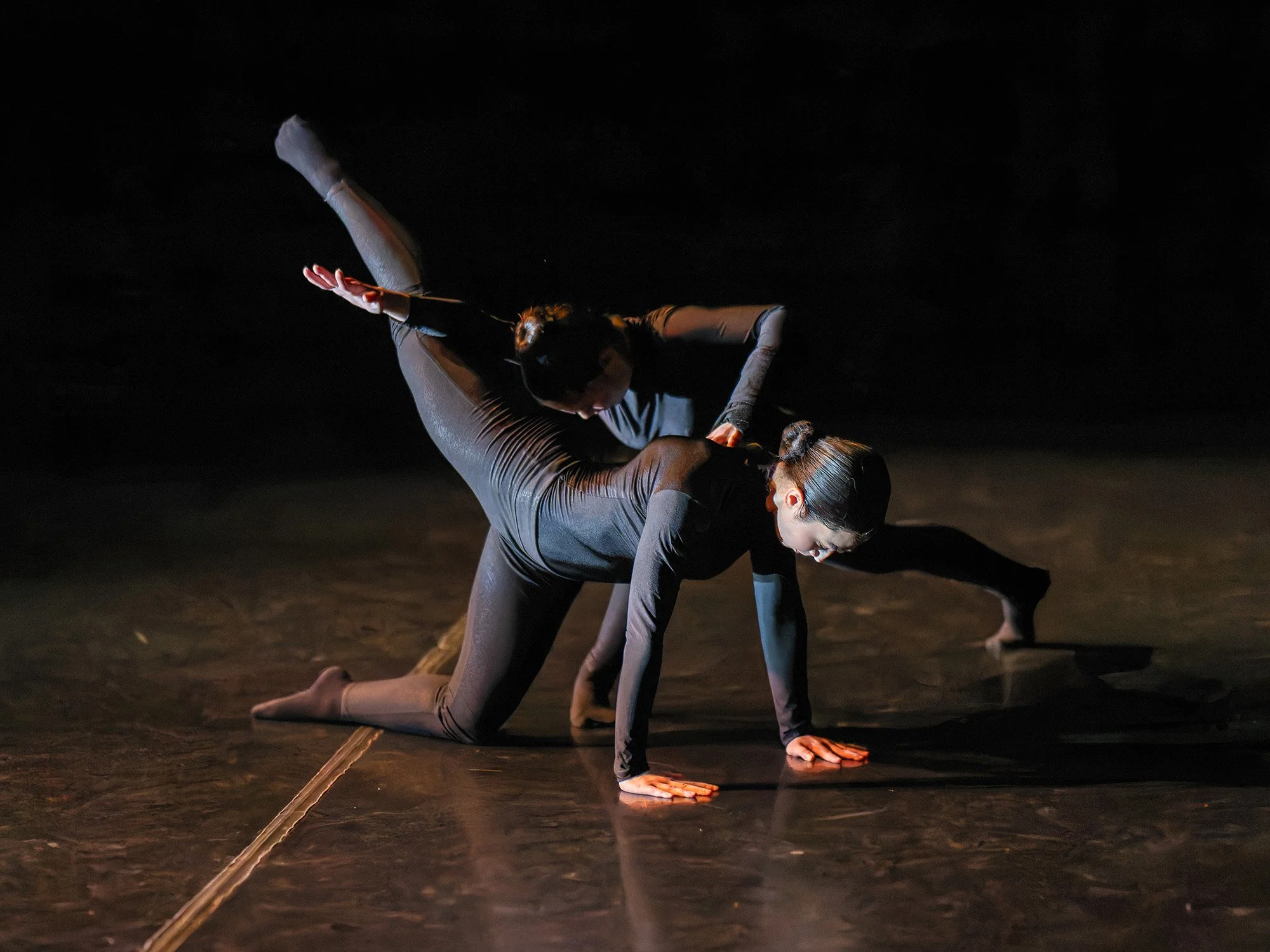 Two dancers in black costumes perform an intricate dance move on a dark stage, with one dancer on hands and knees and the other balancing on one leg while supporting the first dancer.