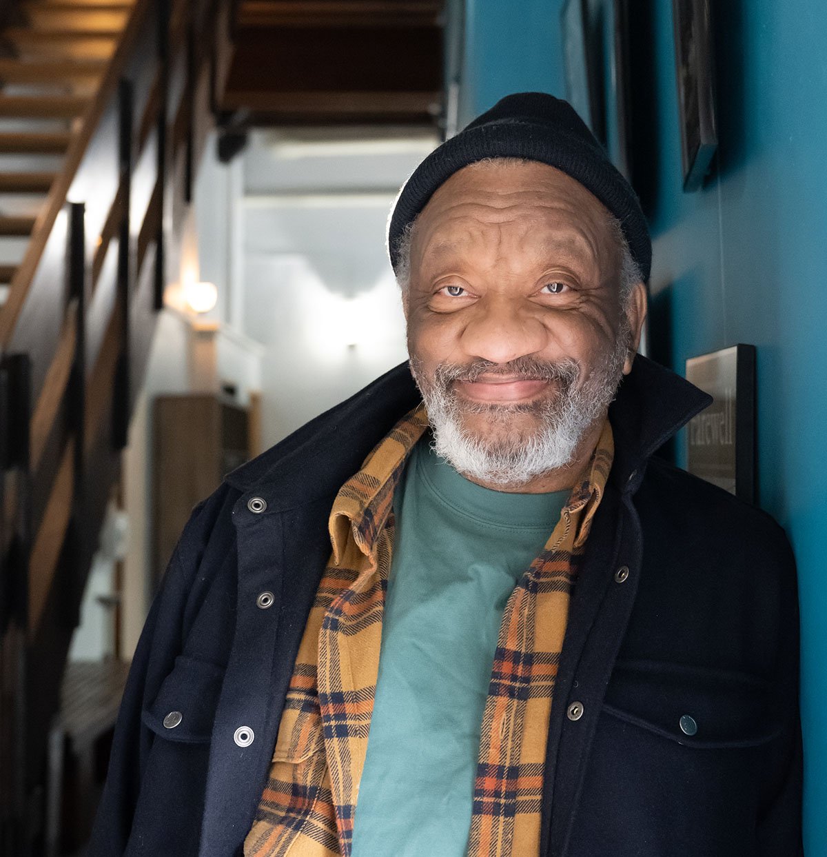 A smiling older man with a beard, wearing a black beanie, green shirt, and plaid jacket, standing indoors against a blue wall.