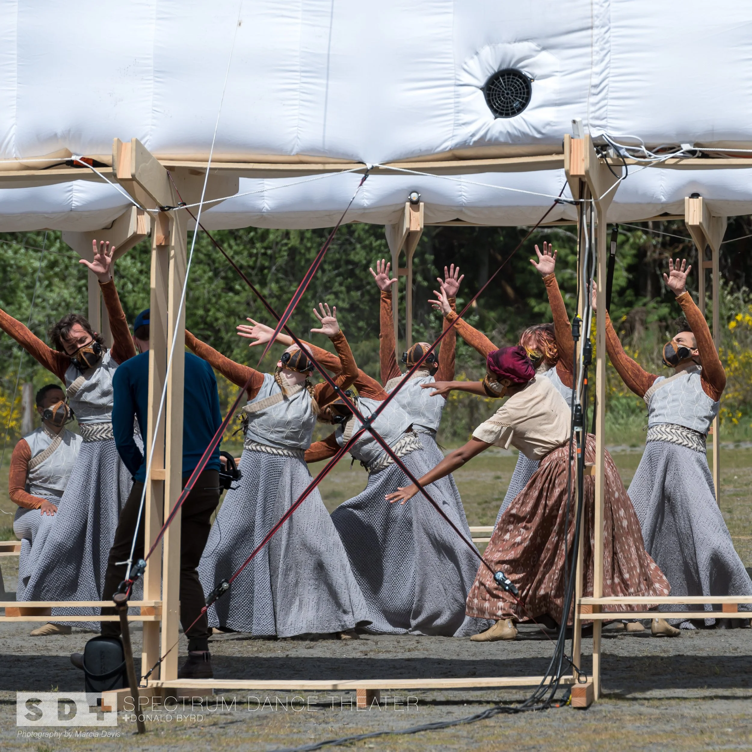 Performers dressed in traditional dresses dancing on an outdoor stage with a wooden frame and a white canopy, with men and women wearing masks and arm raised, surrounded by trees.
