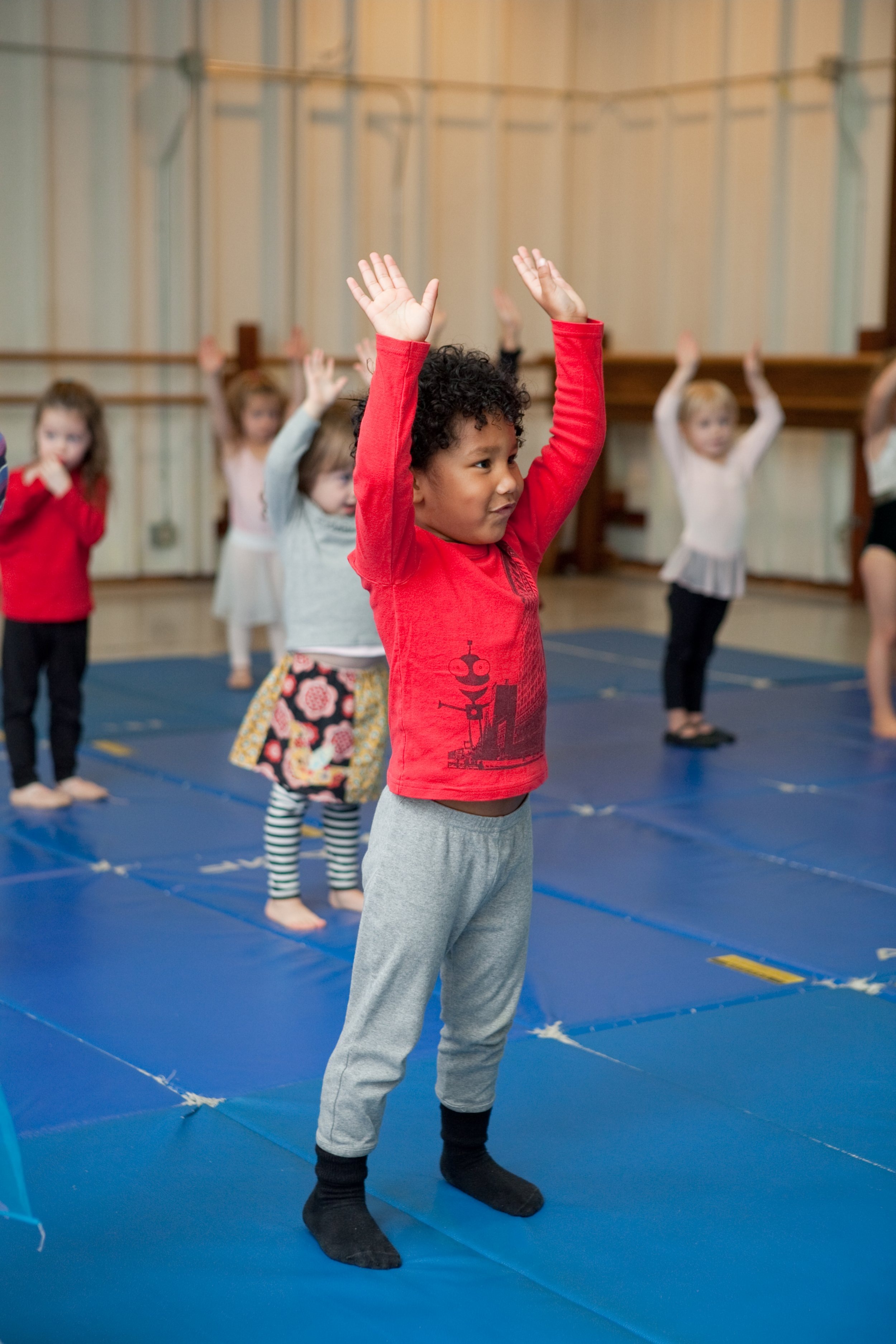Children participating in a gymnastics or dance class, standing on blue mats with their hands raised above their heads.