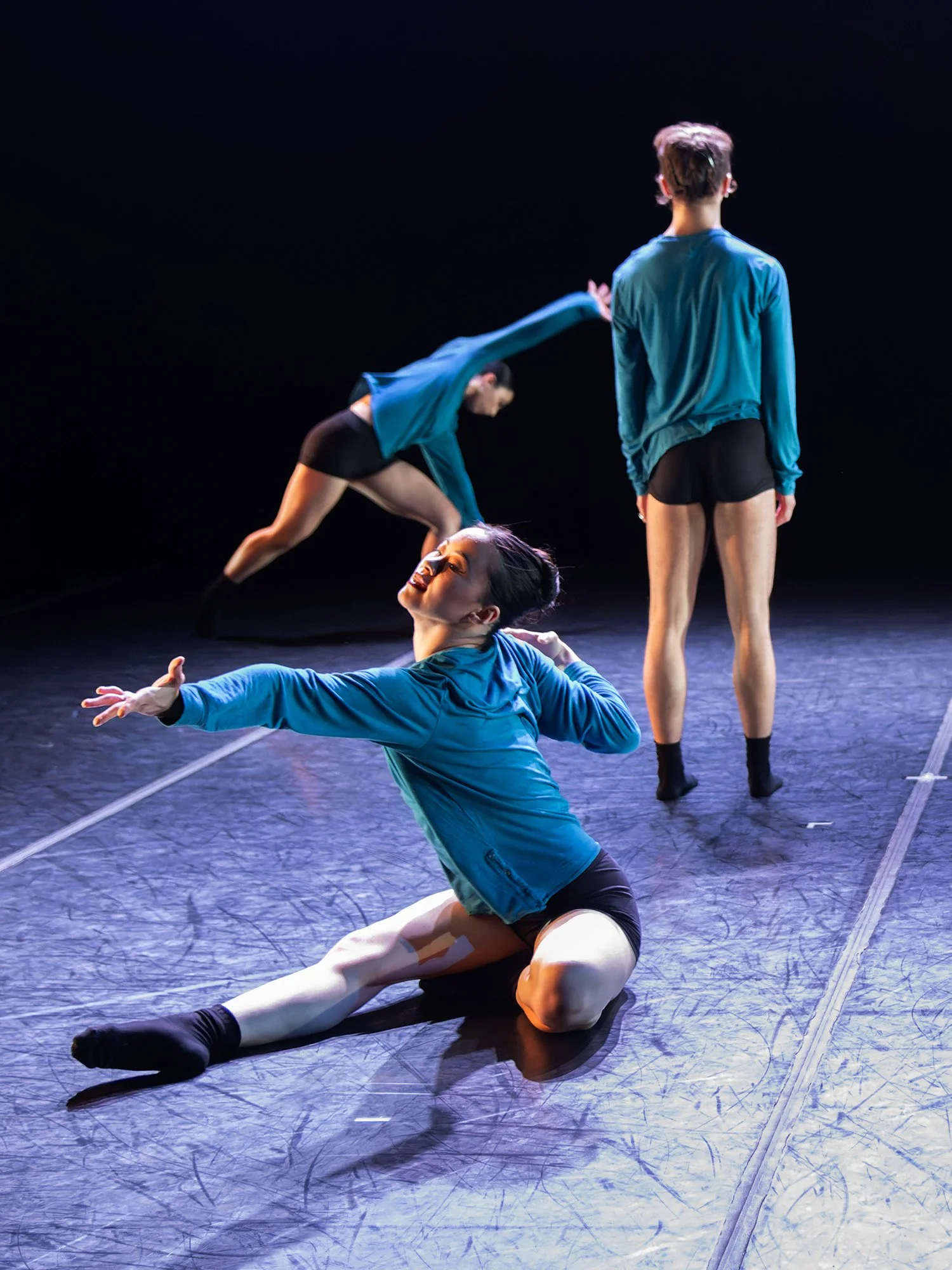 Three dancers performing on stage, with one dancer sitting on the floor in the foreground, one standing in the background, and the third in mid-air, all dressed in blue tops and black shorts or leggings.