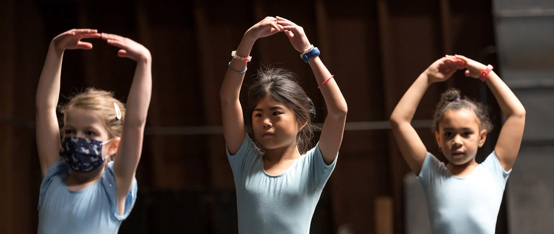 Three young girls practicing ballet or dance with arms raised above their heads, wearing casual dance practice clothing, in a dance studio.