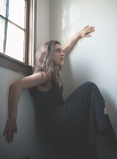 Woman with curly hair sitting on a windowsill with her back against a wall, looking out the window.