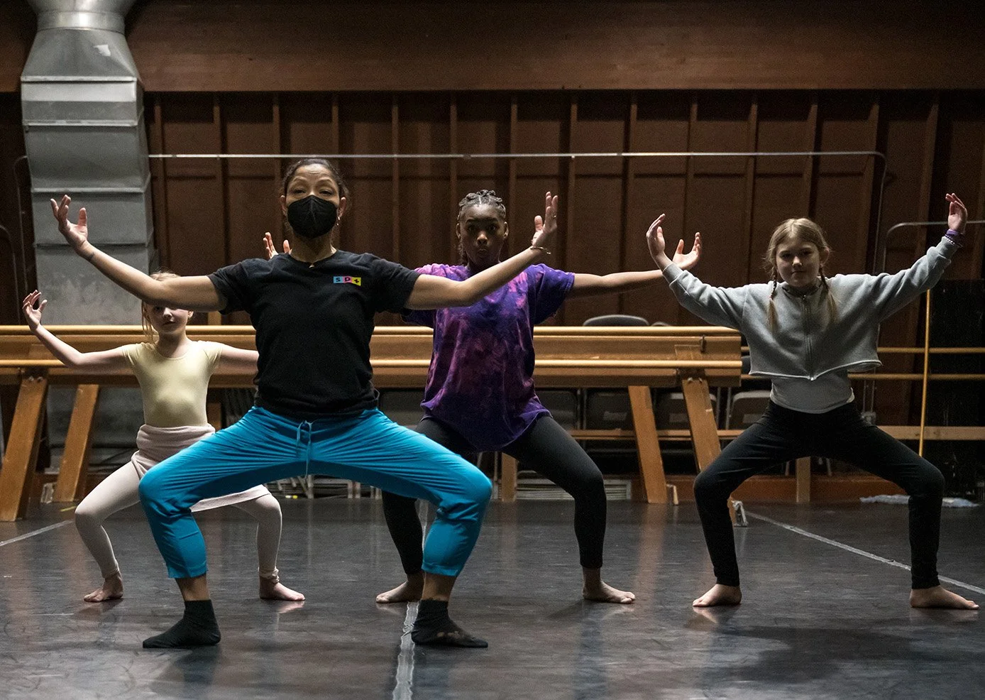 Four children practicing dance or ballet in a studio, with three girls and one adult woman, all in stretching poses with arms extended, on a dark floor with wooden walls and equipment in the background.