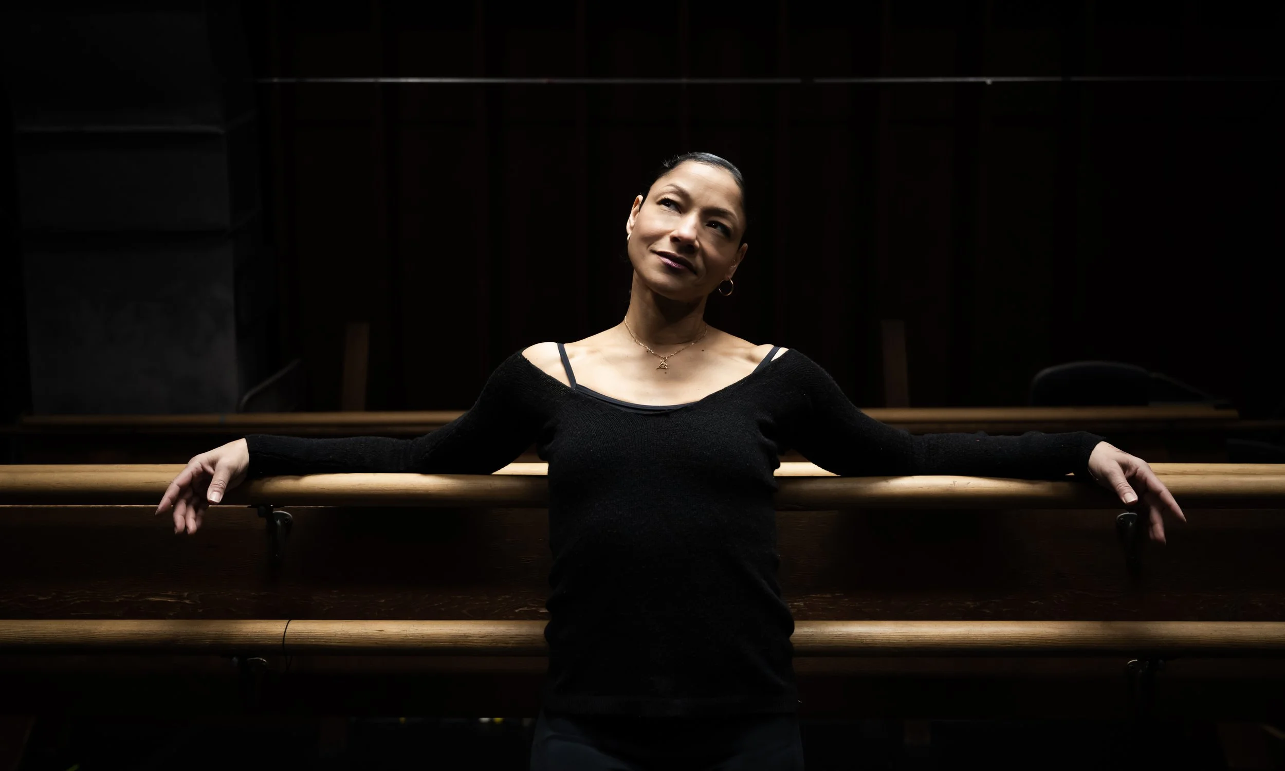 A woman in a black top leaning against a wooden railing with her arms outstretched in a dark room.