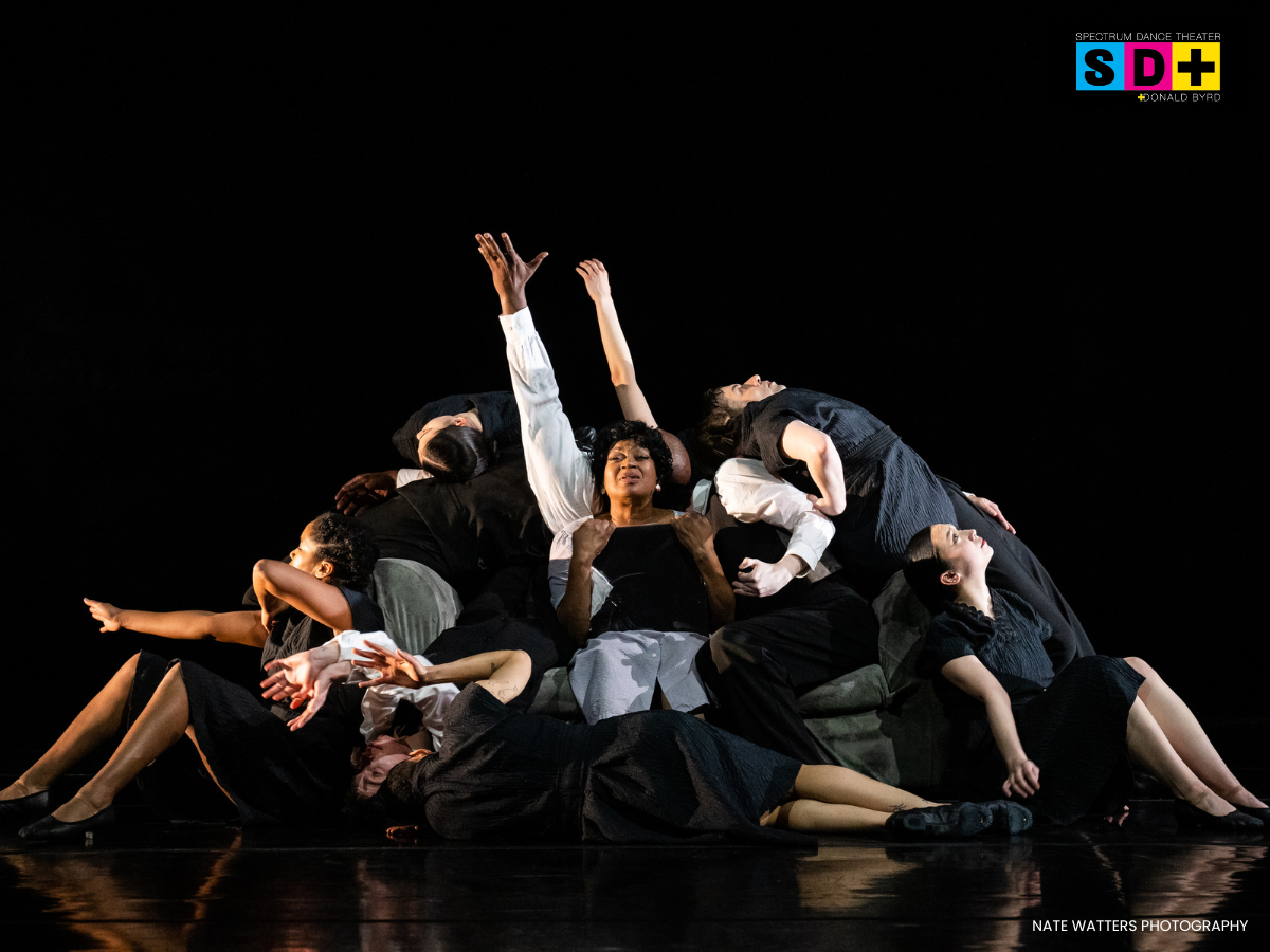 Diverse group of dancers on stage in a theatrical dance performance, posing with expressive gestures and dramatic lighting, with Spectrum Dance Theater logo in the top right corner.