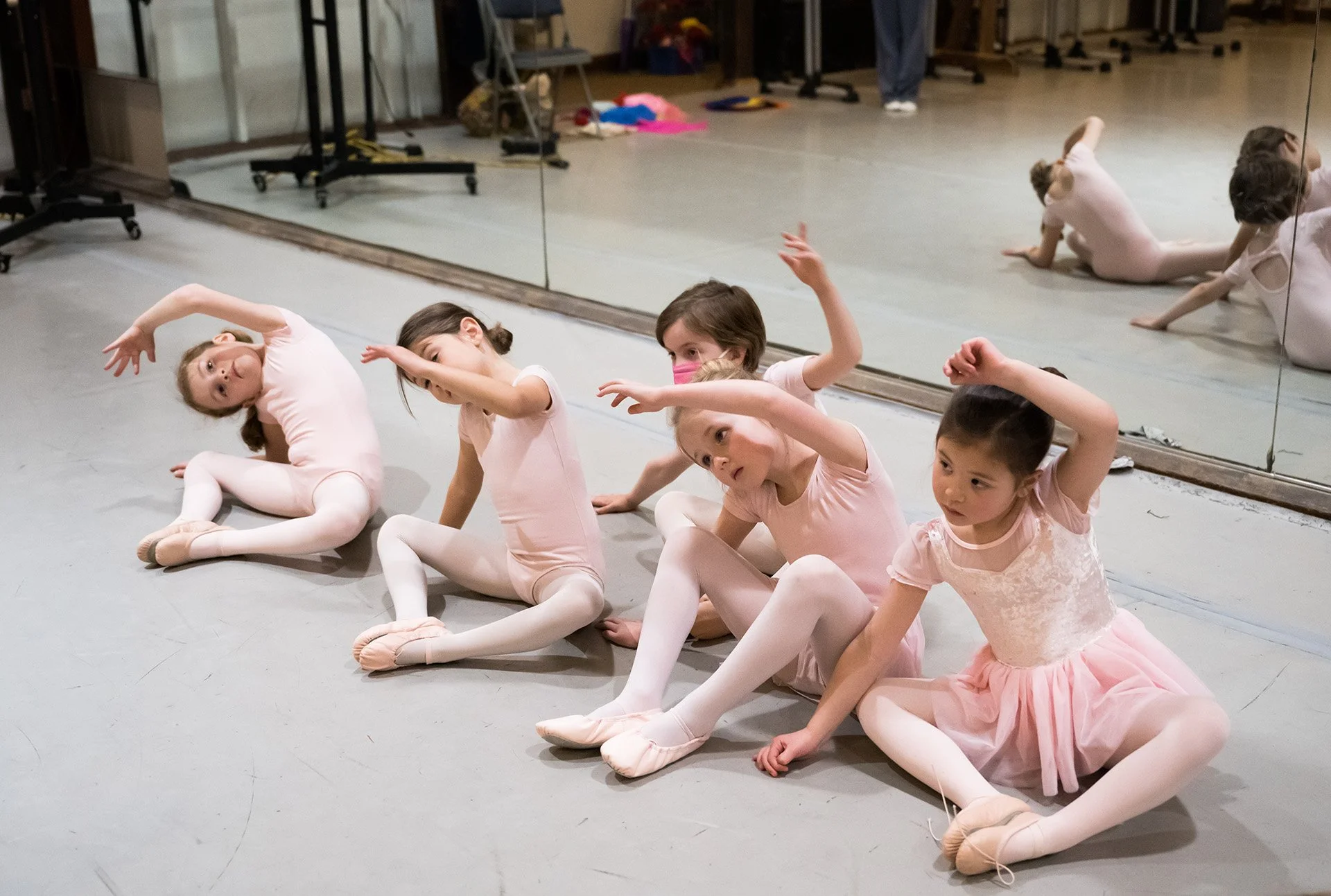 Four young girls in pink ballet outfits sitting on the floor stretching in a dance studio with mirrors.