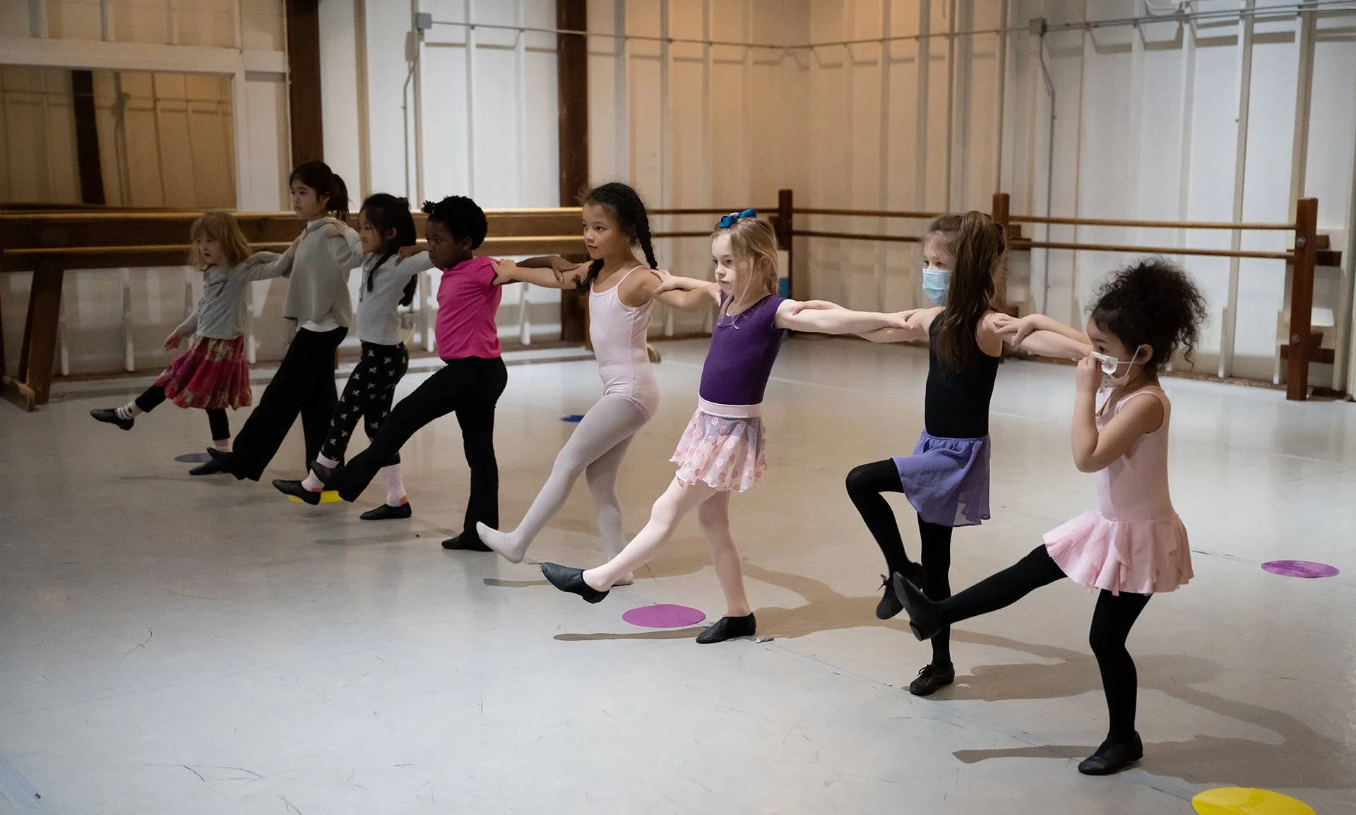 Group of young girls in a ballet class practicing dance moves, some wearing face masks, in dance studio.