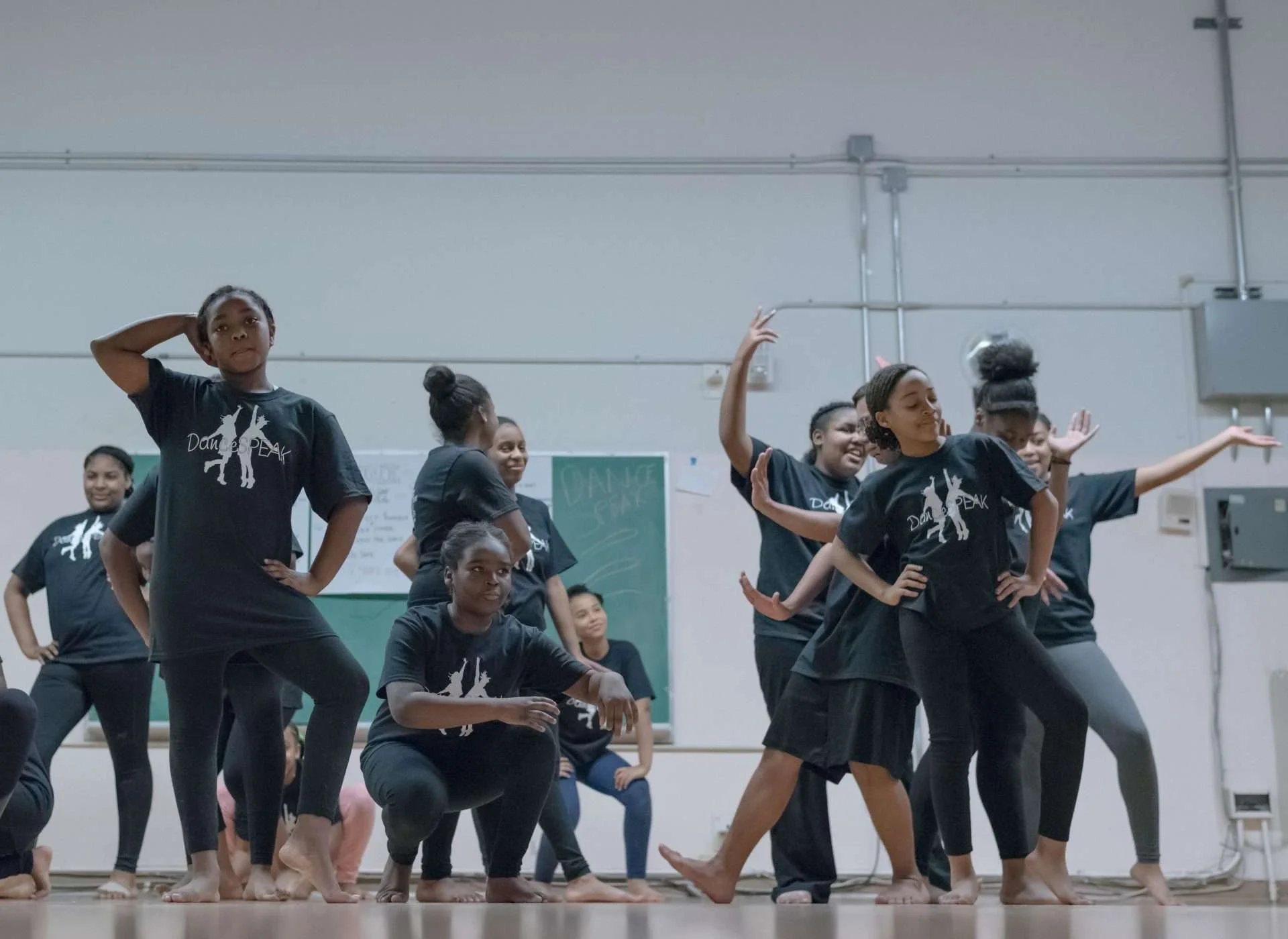 Group of young girls practicing a dance routine in a studio, wearing black t-shirts with a dance logo, some standing with hands on hips, others crouching or dancing.