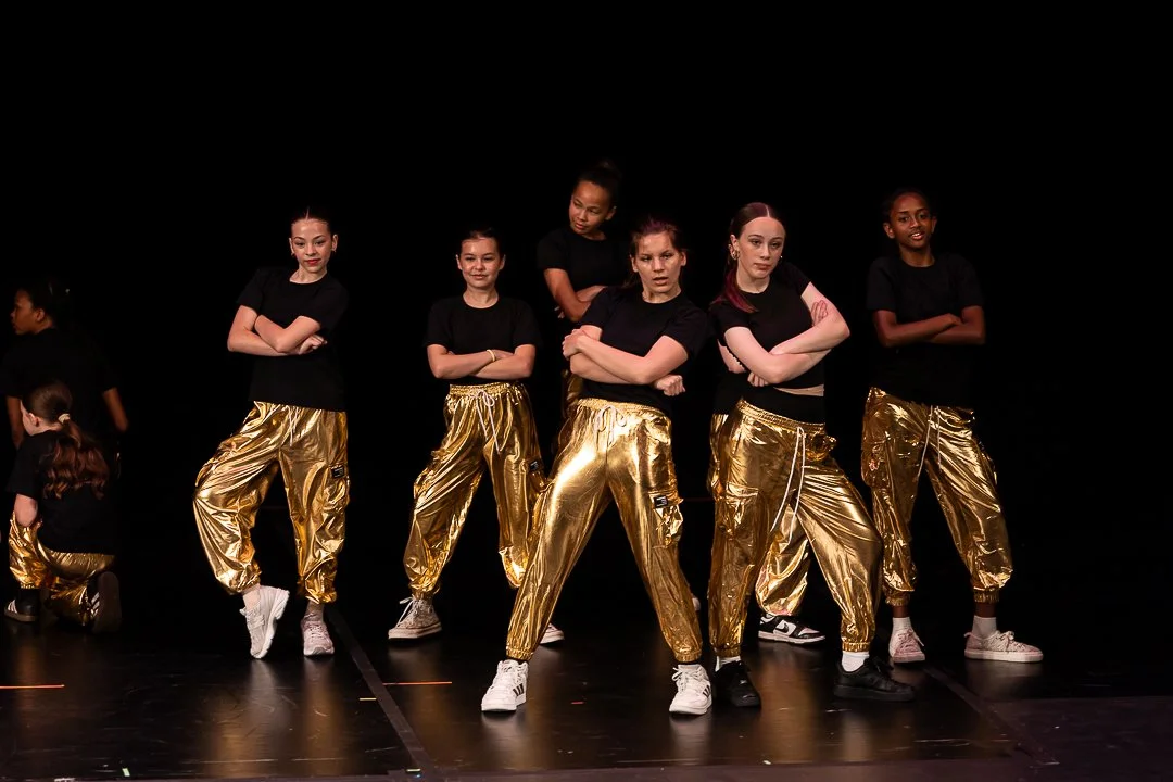 Group of young women performing a dance on stage, wearing black t-shirts and shiny gold pants.