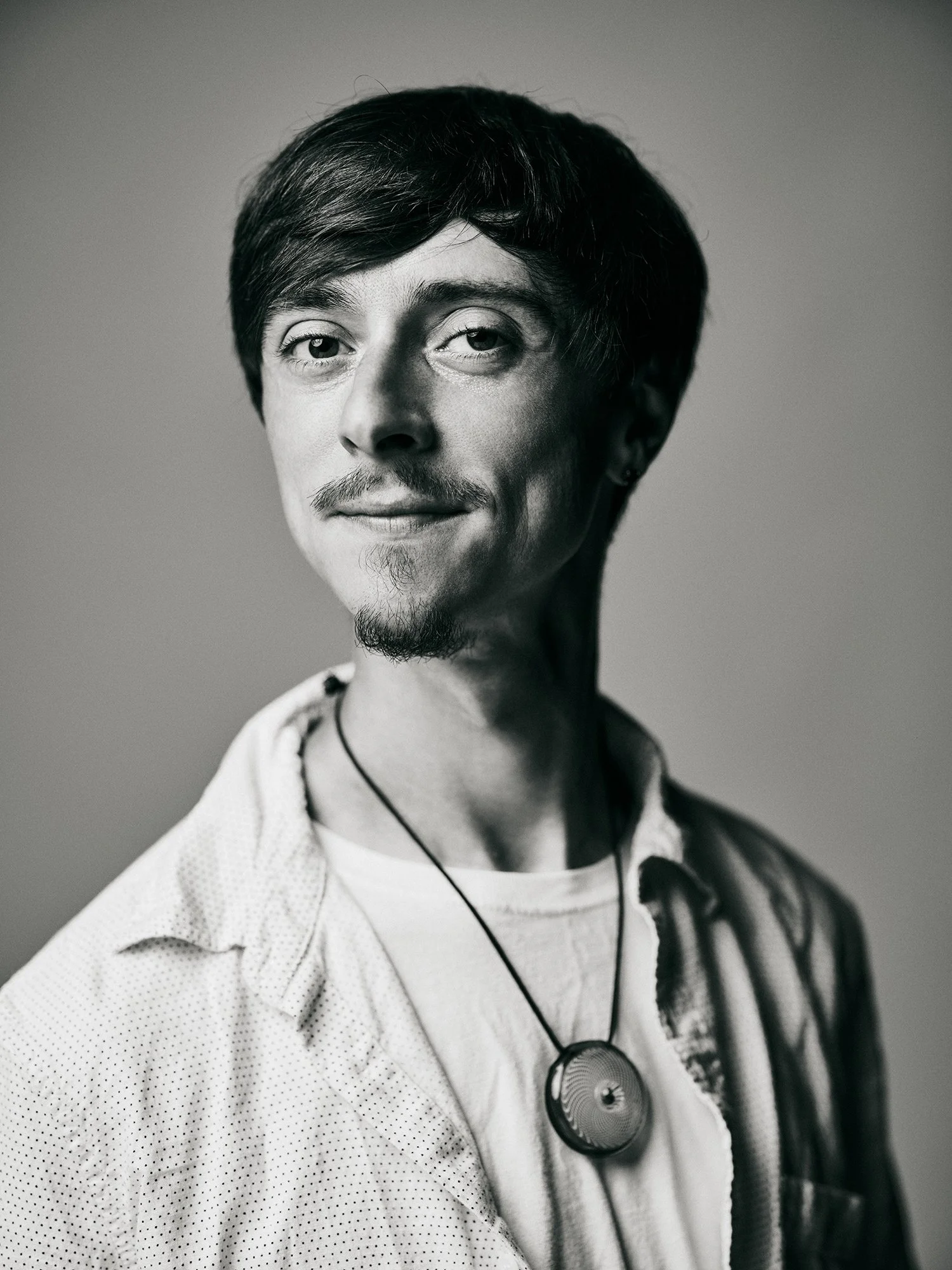 Black and white portrait of a man with dark hair, mustache, and goatee, wearing a white shirt and a pendant necklace, looking confidently at the camera.