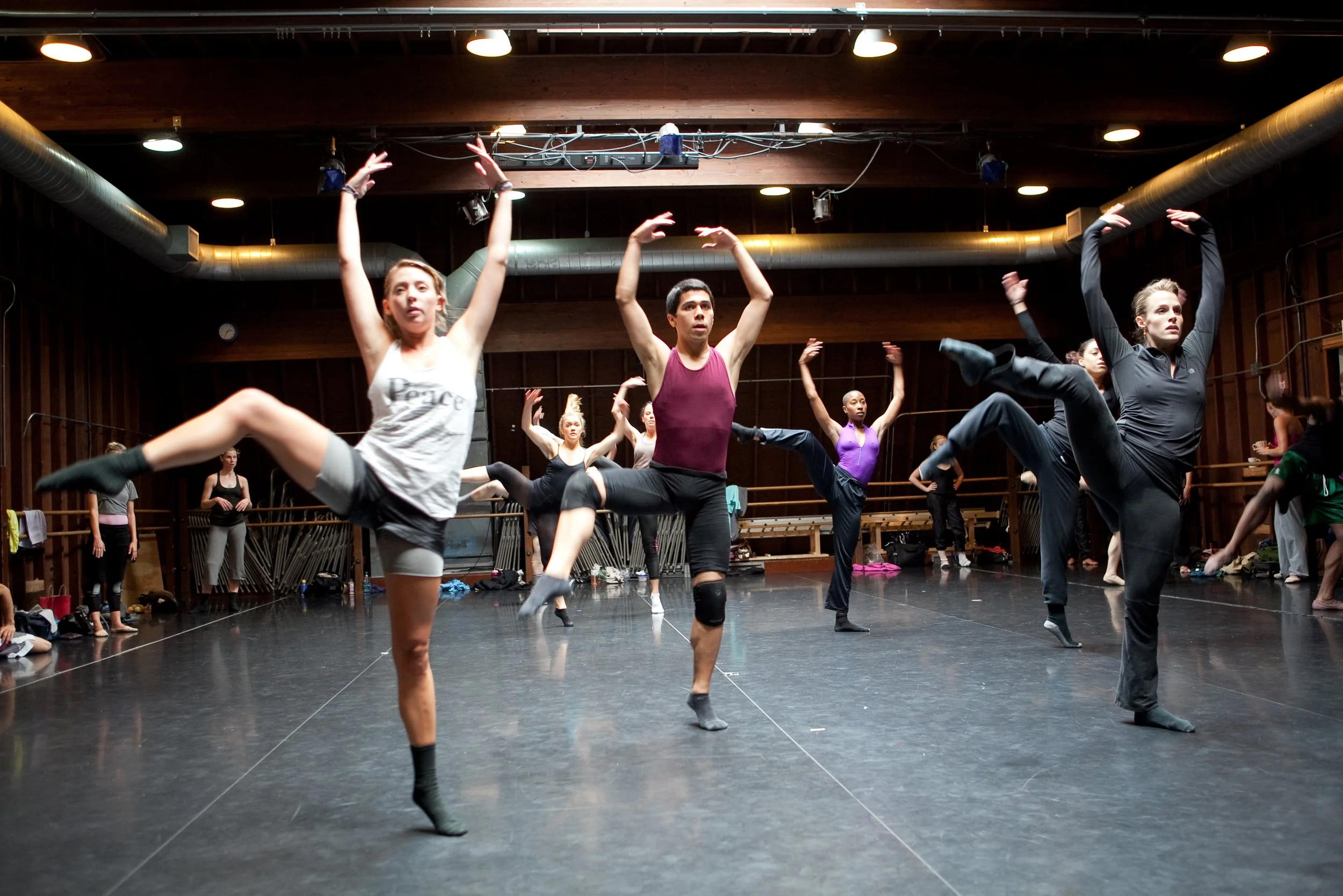 Dance students practicing in a dance studio with a mirror and wooden walls, some stretching and warming up.