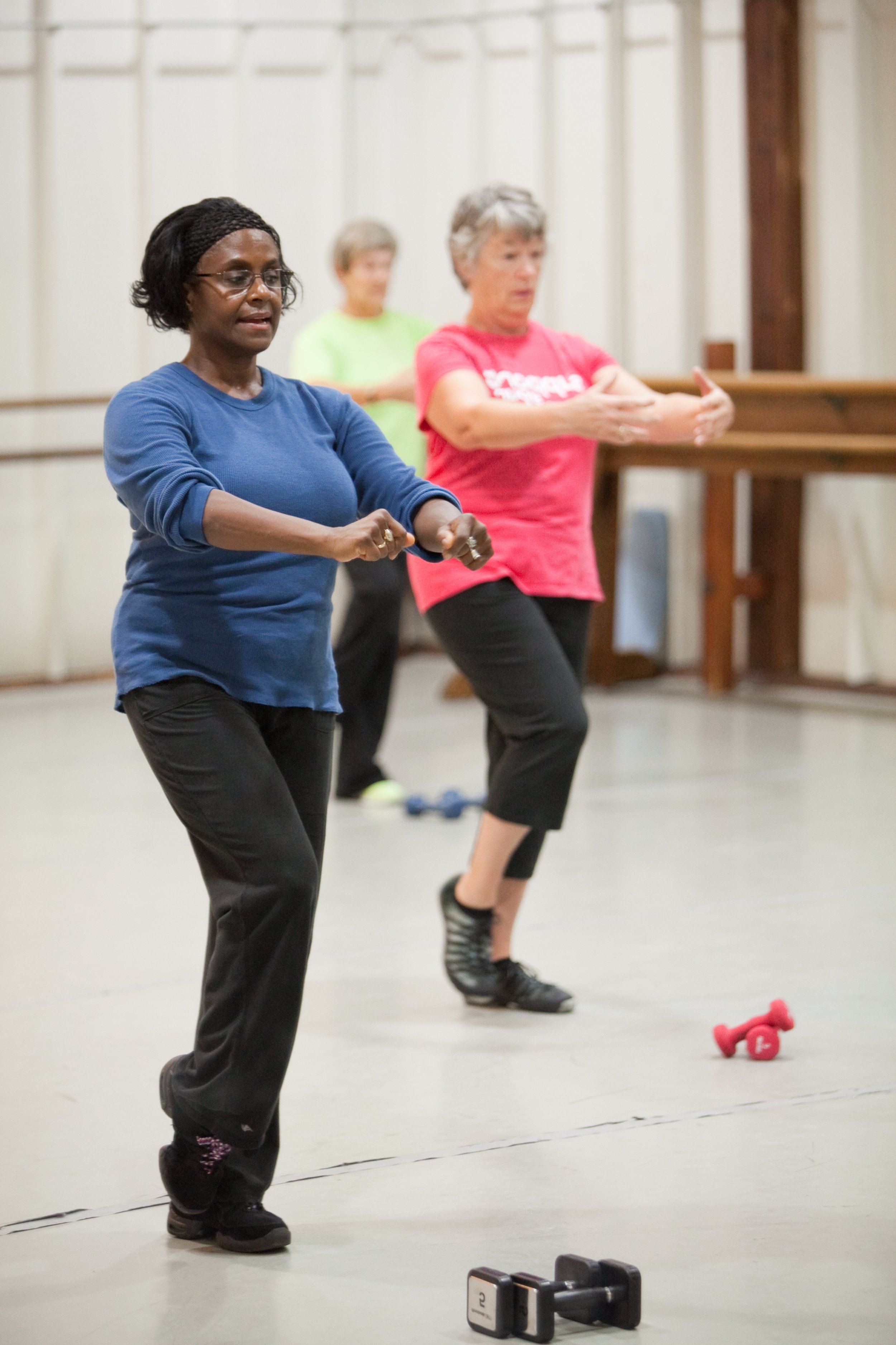 Three women exercising in a fitness class with small weights and dumbbells, practicing arm movements.
