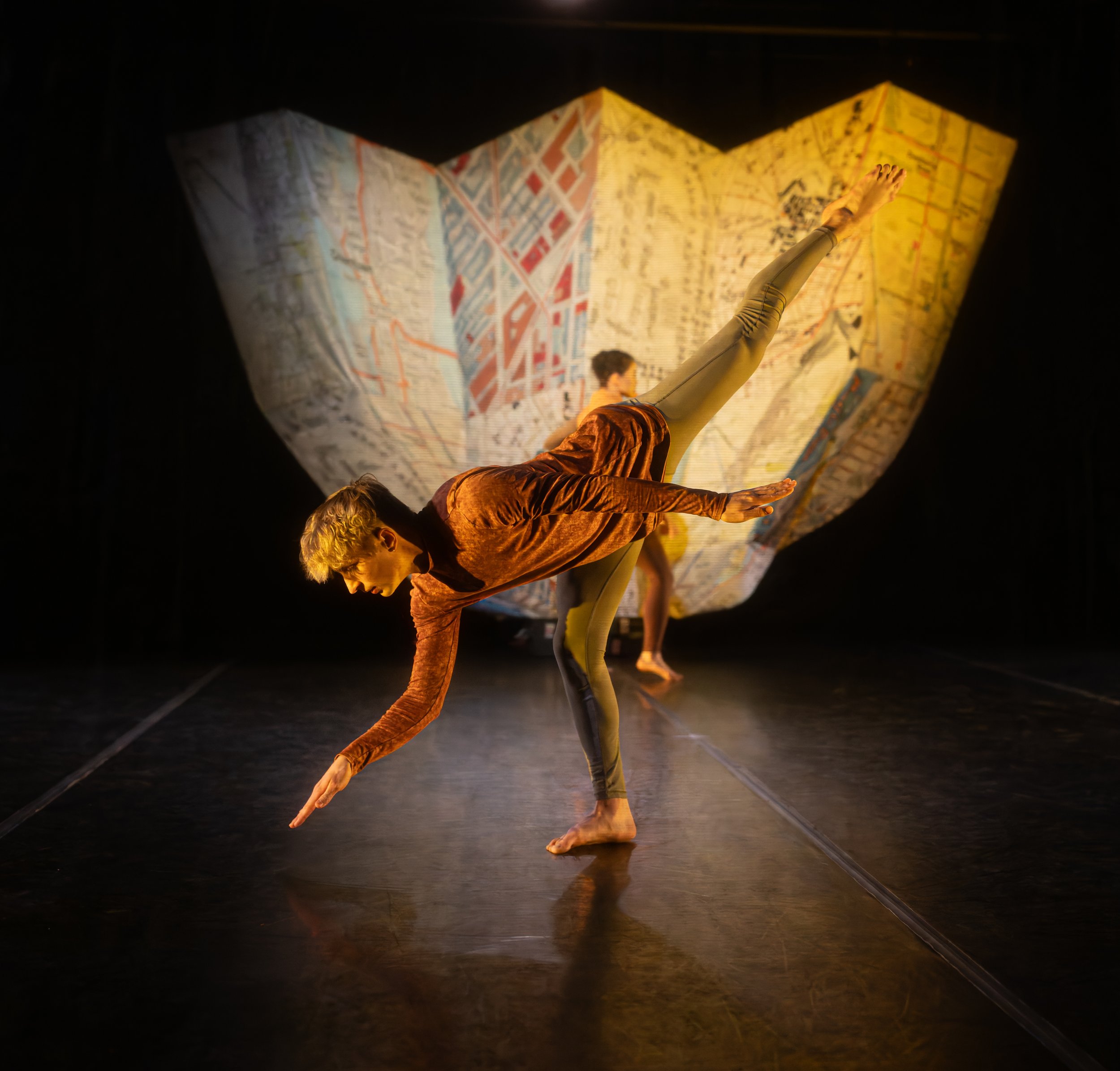 Two performers on stage, one balancing on one leg with the other leg extended behind and arms parallel to the ground, in front of a large backdrop resembling a map.