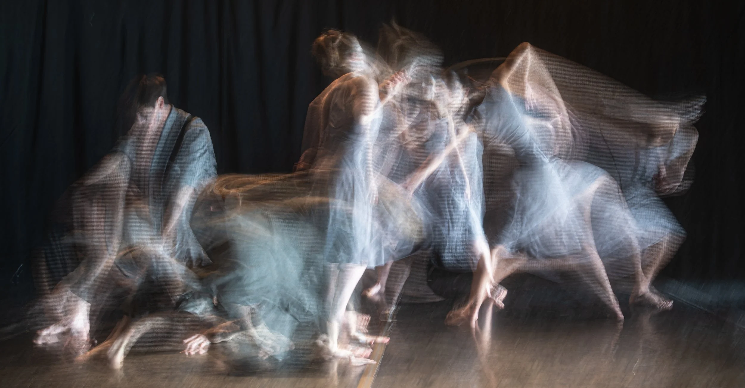 Multiple people in motion blur, performing on stage with a dark curtain background, creating a layered, abstract effect.