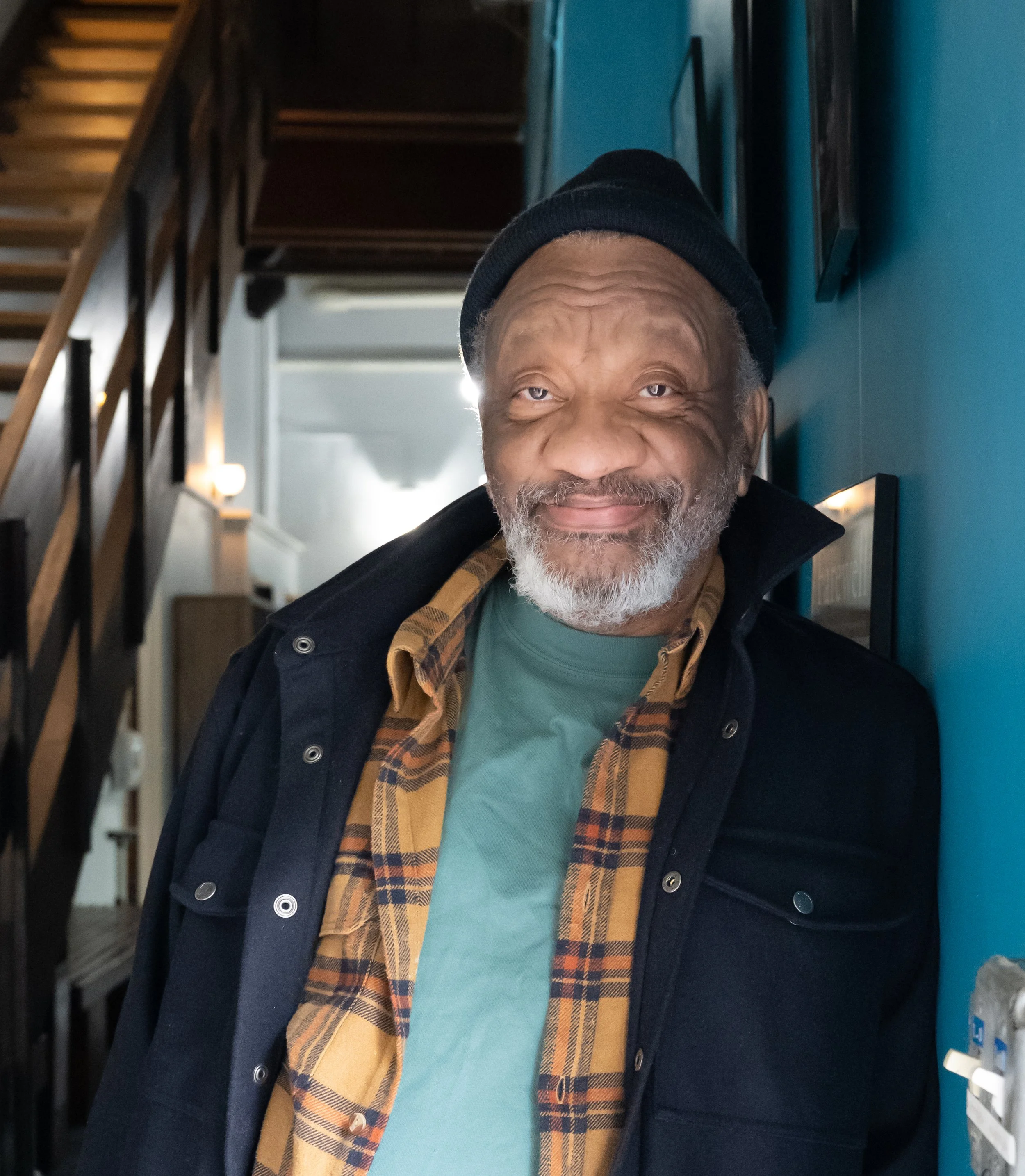 Older man with gray beard smiling, wearing a black beanie and layered clothing, standing against a blue wall in a modern interior.