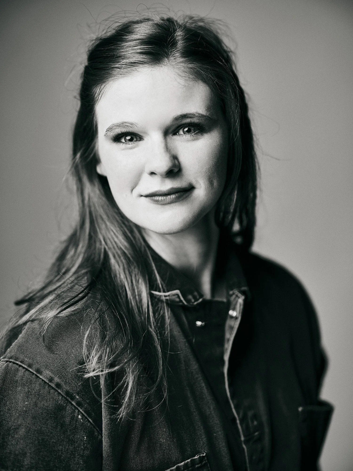 Black and white portrait of a woman with long hair, wearing a denim jacket, looking at the camera with a slight smile.
