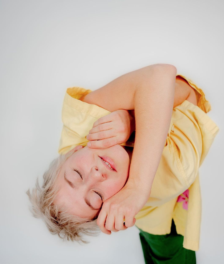 A woman with short, light blonde hair lying on a white surface, wearing a yellow shirt, with her eyes closed and hand near her face.