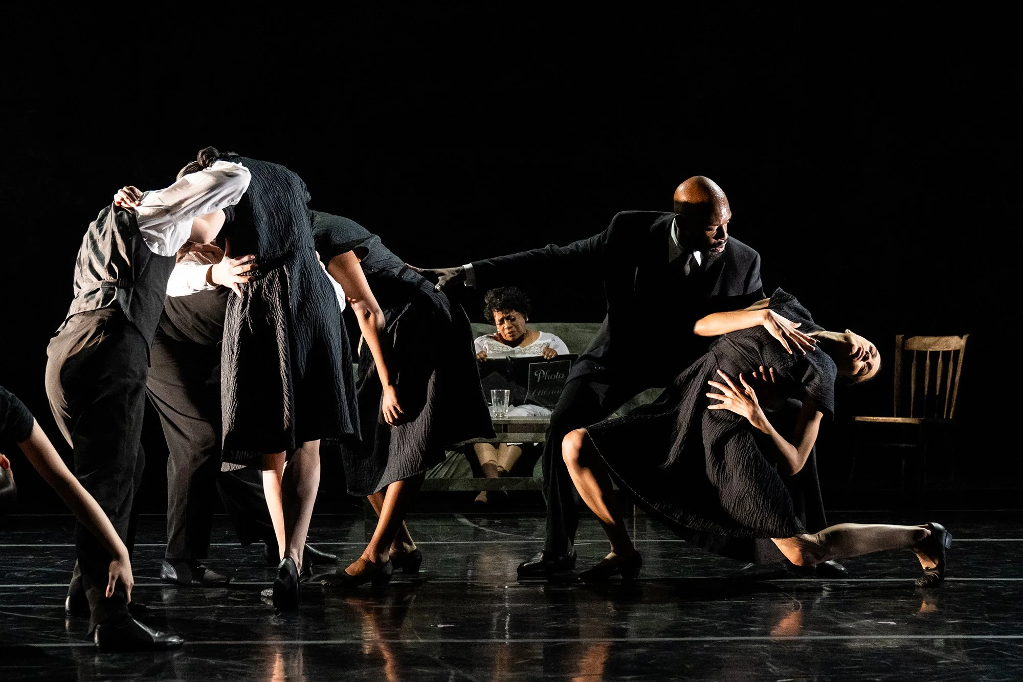 Actors performing on stage with dramatic lighting, some leaning or kneeling, in a theatrical scene.