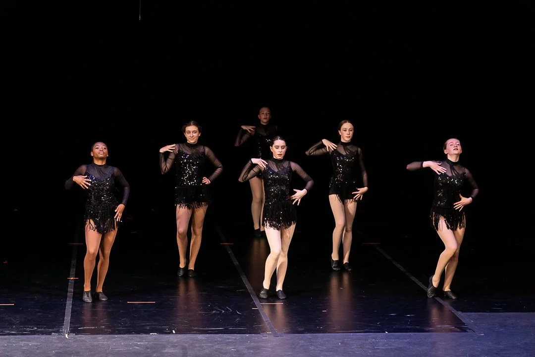 Six female dancers in black costumes performing on stage with a dark background.