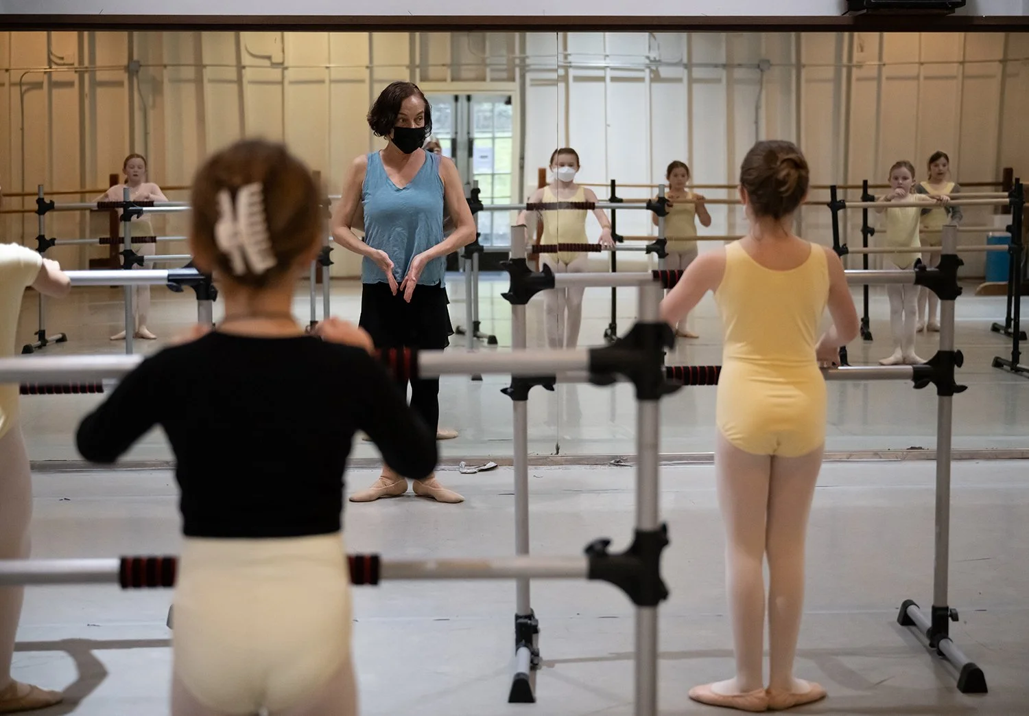 Ballet class with young girls practicing at barre while an instructor gives instructions in a dance studio.