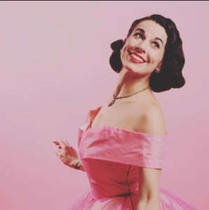 A smiling woman with dark hair styled in vintage waves, wearing a pink off-the-shoulder dress against a pink background.