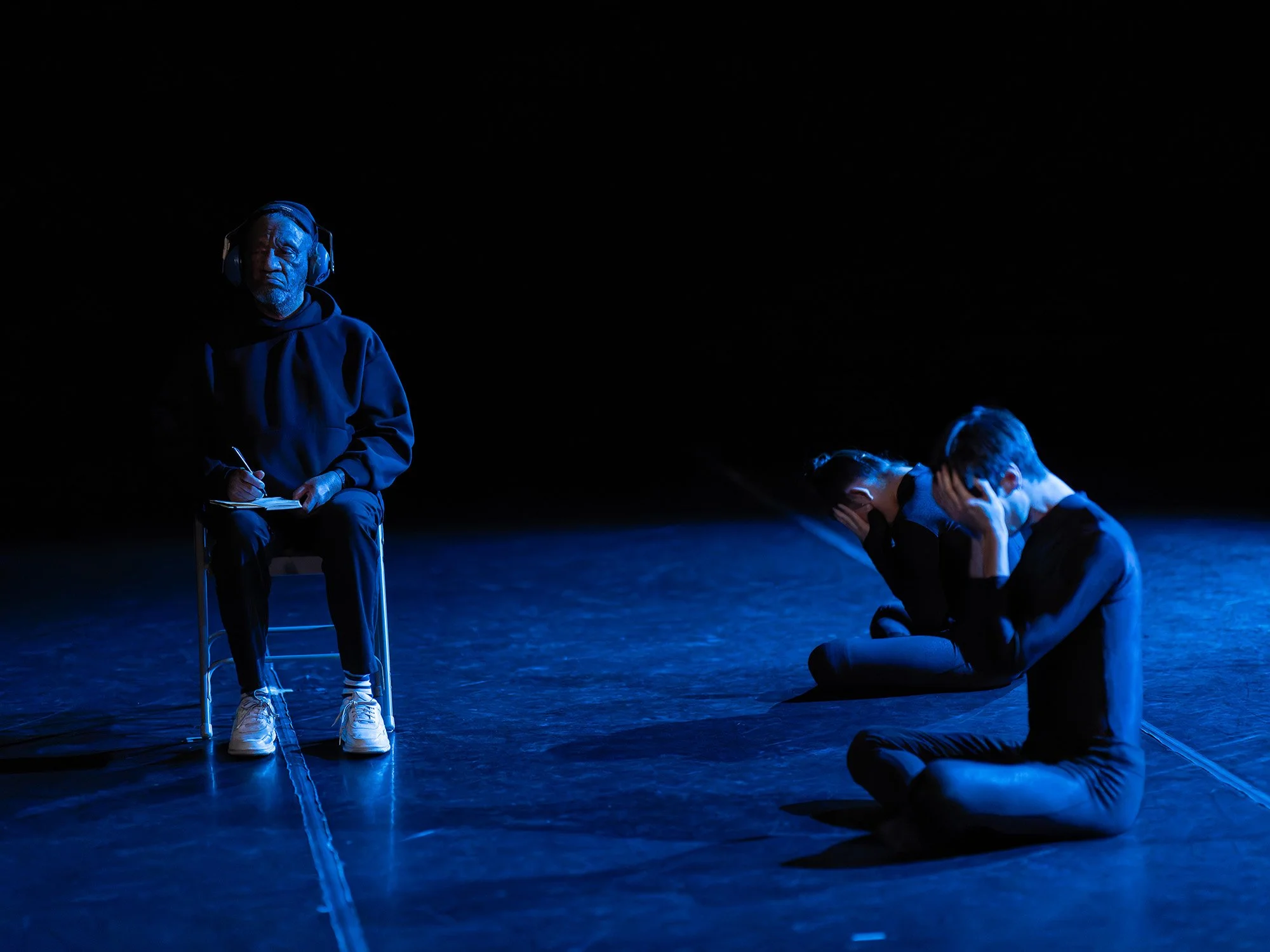 Three performers on a dark stage illuminated by blue light, two sitting on the floor with heads bowed and hands covering their faces, one sitting on a chair wearing headphones and writing in a notebook.