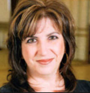 A woman with shoulder-length brown hair and bangs, smiling, in an indoor setting with yellow curtains in the background.