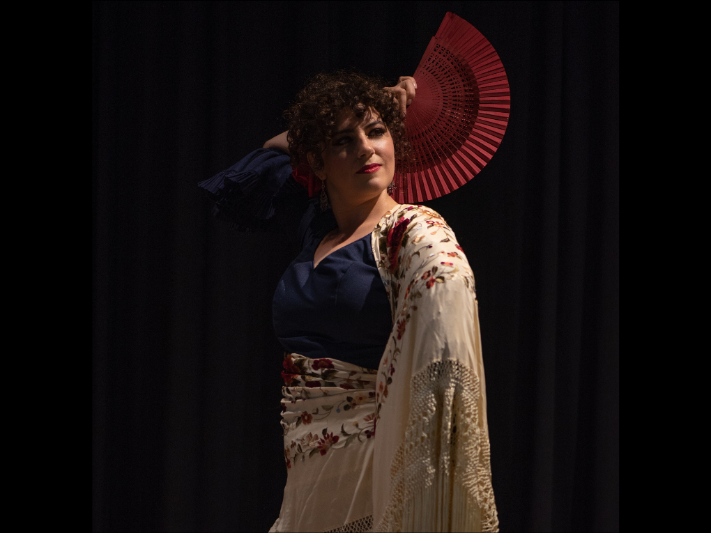 A woman with curly hair dressed in a traditional flamenco outfit, holding a red fan near her head, standing against a dark curtain backdrop.
