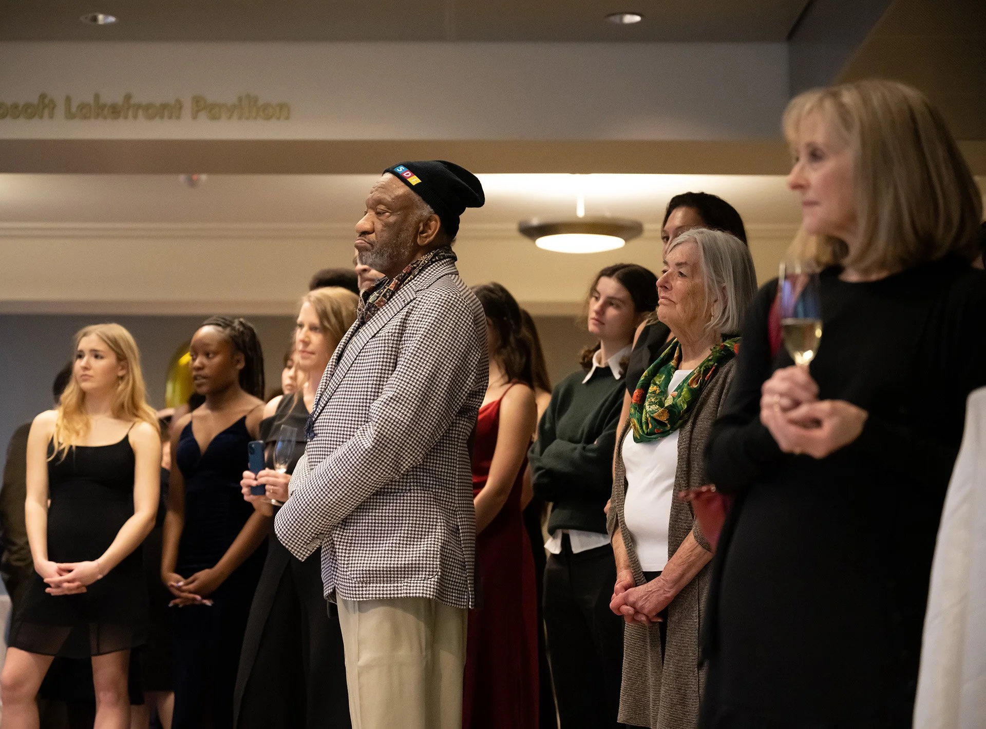 People standing in a room, listening attentively at what appears to be a formal event or gathering, some holding drinks.