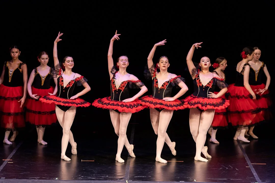 A group of young ballet dancers performing on stage in black and red costumes, with some dancers in the background and a dark stage backdrop.