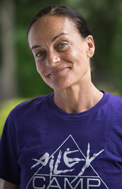 Close-up of a woman with dark hair pulled back, wearing a purple t-shirt with the text 'ALEX CAMP' and a geometric logo, standing outdoors with a blurred green background.