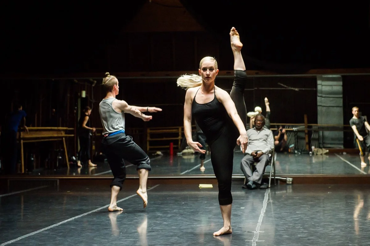 Ballet dancers practicing on a dance studio stage with mirrored wall, instructor seated, and other dancers in the background.
