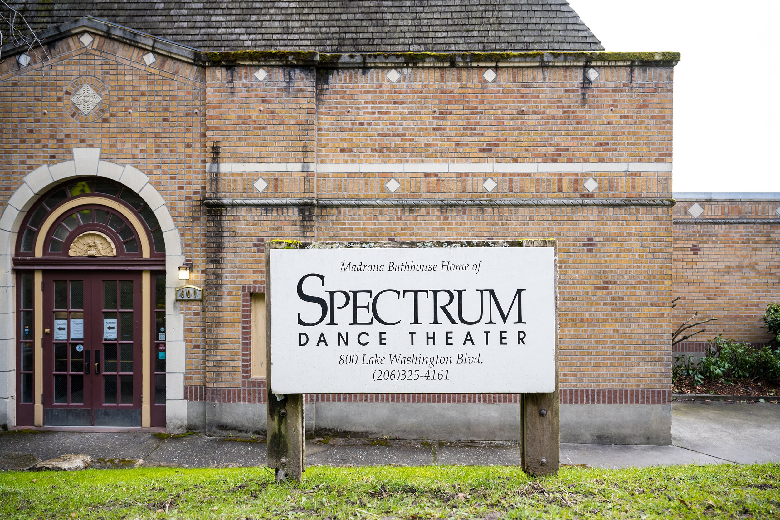A brick building with a sign indicating it is the Spectrum Dance Theater, located at 800 Lake Washington Blvd, Washington, with a phone number (206) 325-4161. The entrance has a maroon door with glass panes framed in beige, and there is a small outdoor lamp beside the door.