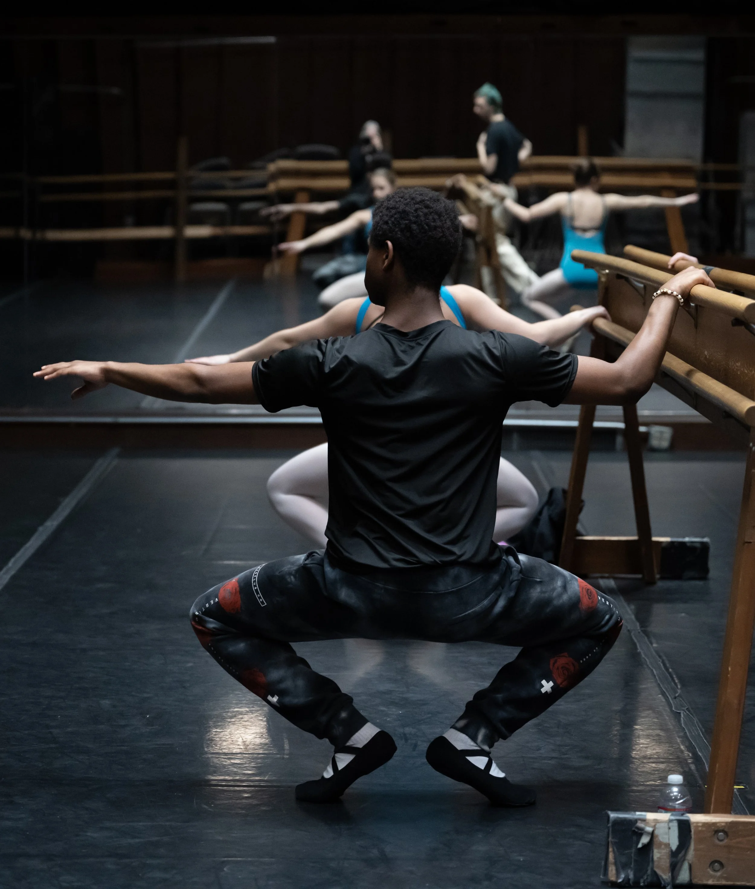 Ballet dancers practicing in a dance studio, with a teacher supervising, one dancer in the foreground squatting with arms extended to the sides.