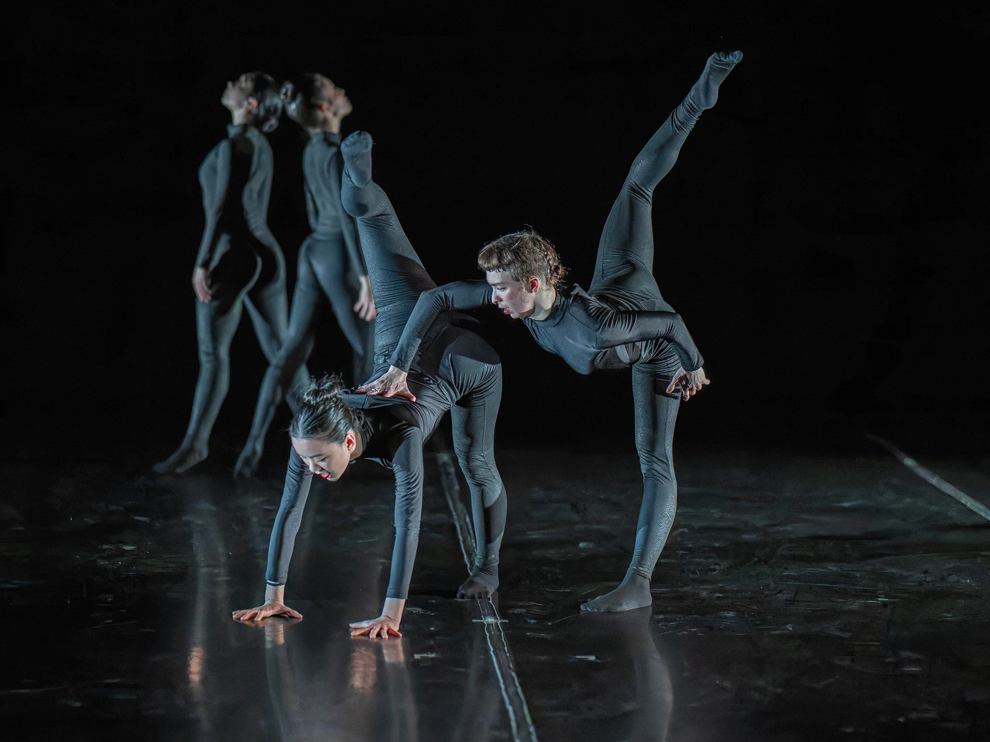 Four dancers in black costumes performing a contemporary dance routine on stage, with two dancers in the background in a standing pose and two in the foreground forming an acrobatic duo with one balancing on the other's back.