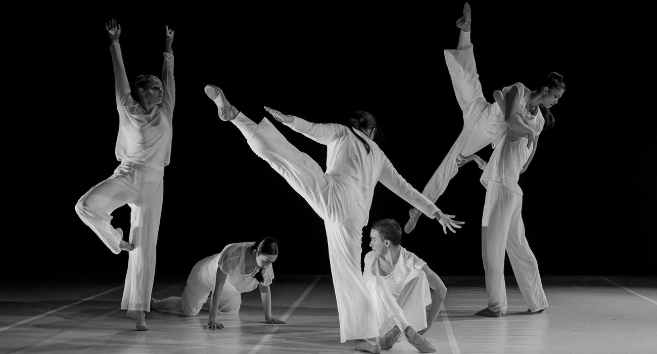 Six dancers in white clothing performing a contemporary dance routine on a dark stage, with some dancers standing, some kneeling, and one doing a high kick.