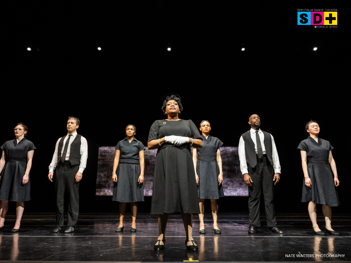 A group of seven performers standing on stage during a theater production, dressed in black and white attire, with a woman in the center wearing white gloves. The stage has black flooring, and a black background with some lighting. The logo for Spect