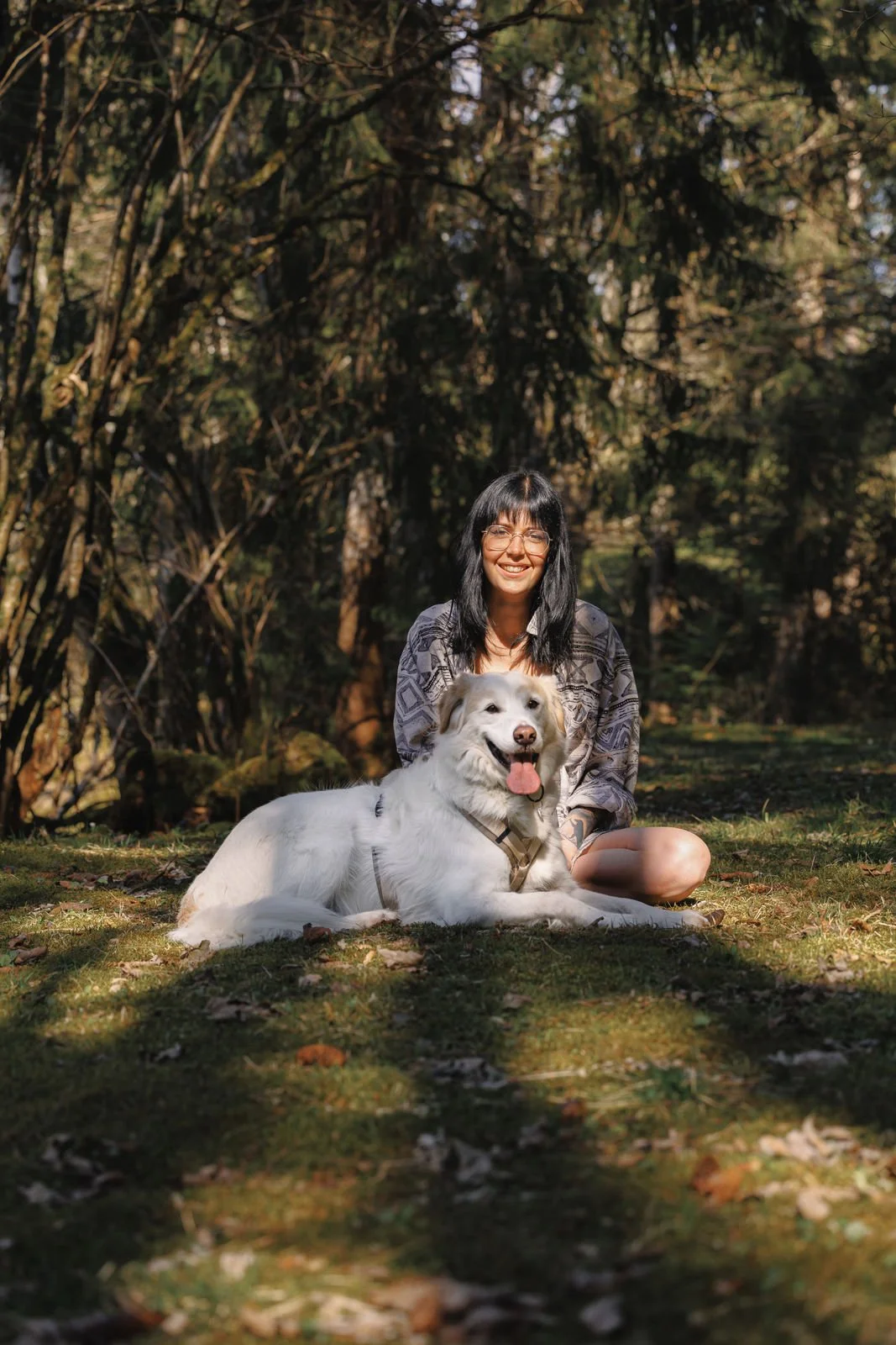 Frau sitzt mit einem lächelnden weißen Hund auf Gras in einem Wald.
