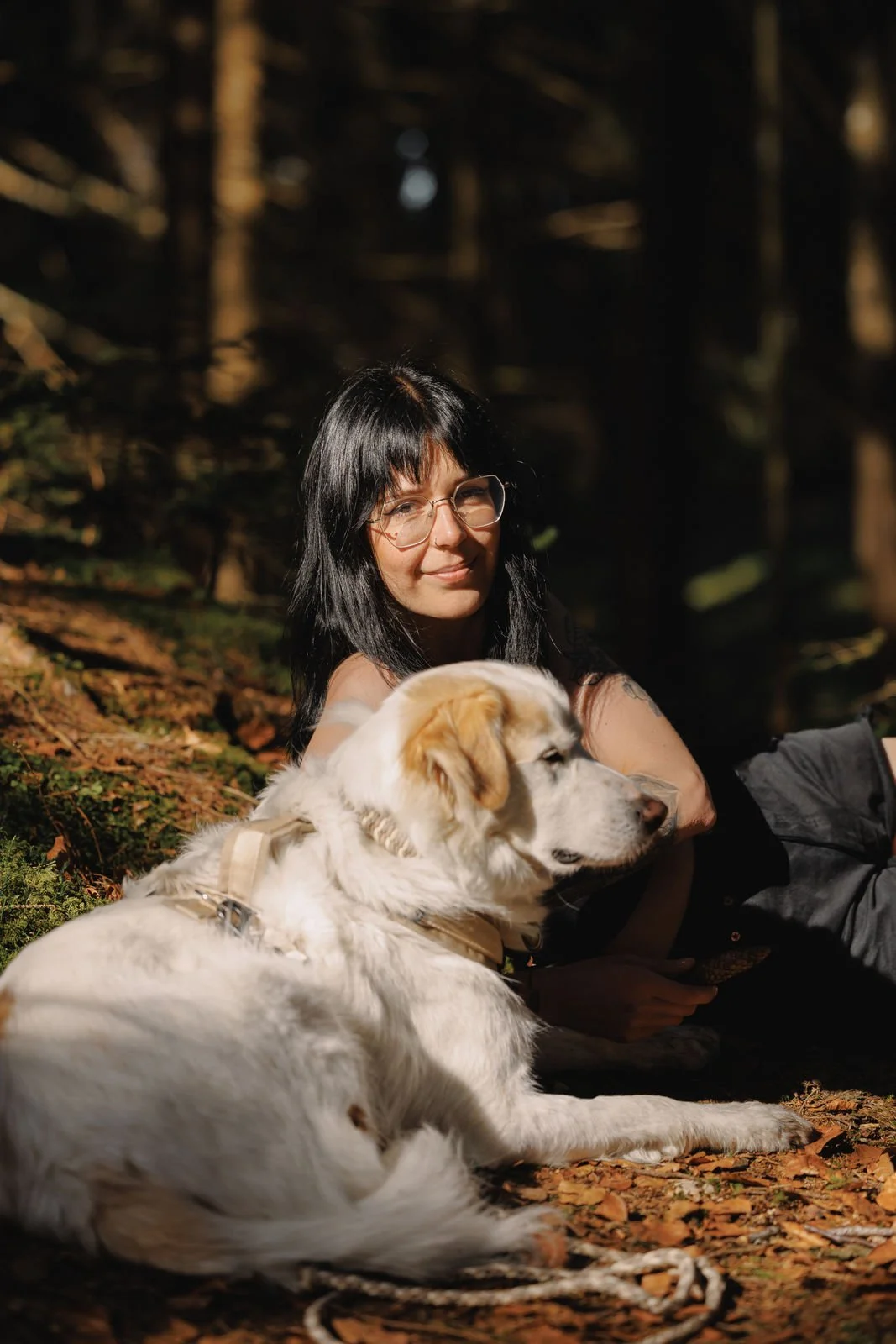 Eine Person mit langen dunklen Haaren und Brille sitzt im Wald im Sonnenschein neben einem großen weißen Hund.