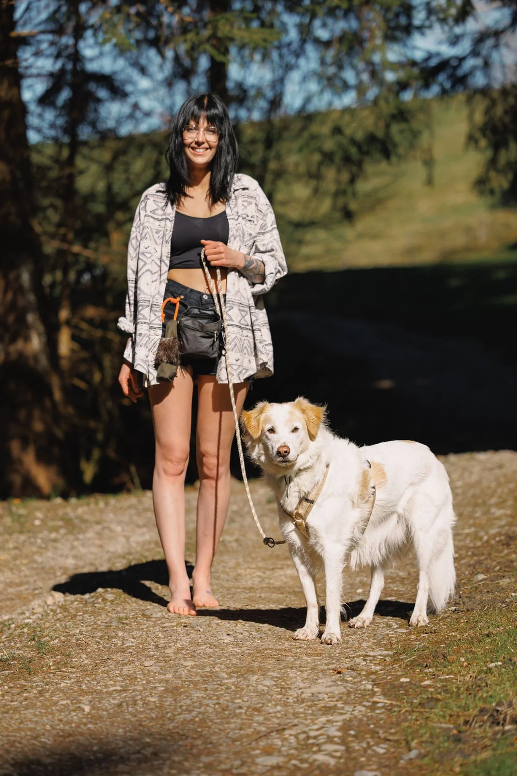 Eine Frau steht draußen auf einem Weg mit einem Hund an der Leine. Die Sonne scheint, und sie trägt sommerliche Kleidung. Im Hintergrund sind Bäume und ein grüner Hügel zu sehen.