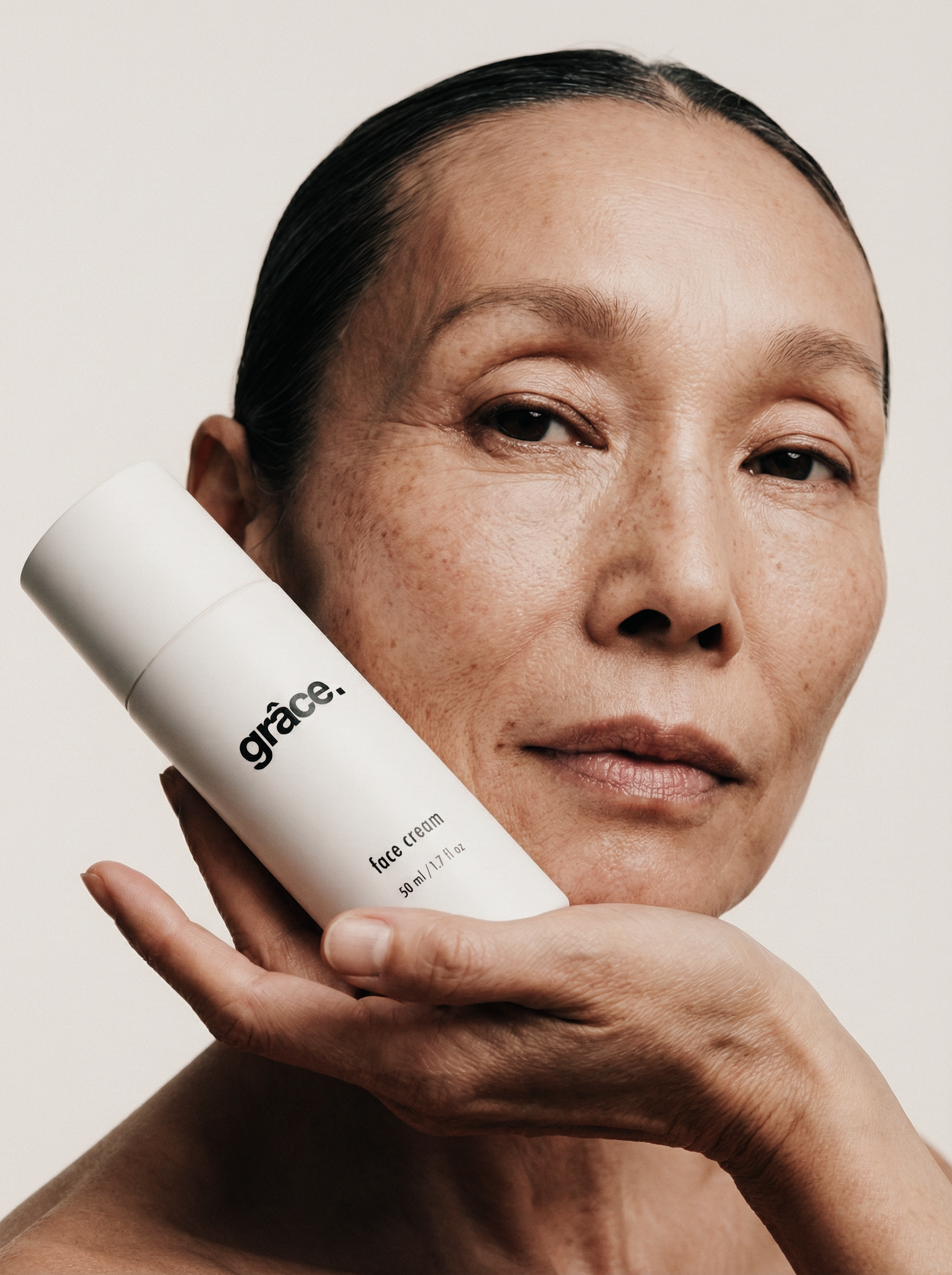 Close-up of a mature woman with fair skin and dark hair, holding a white bottle labeled 'grâce face' near her face, with a neutral background.