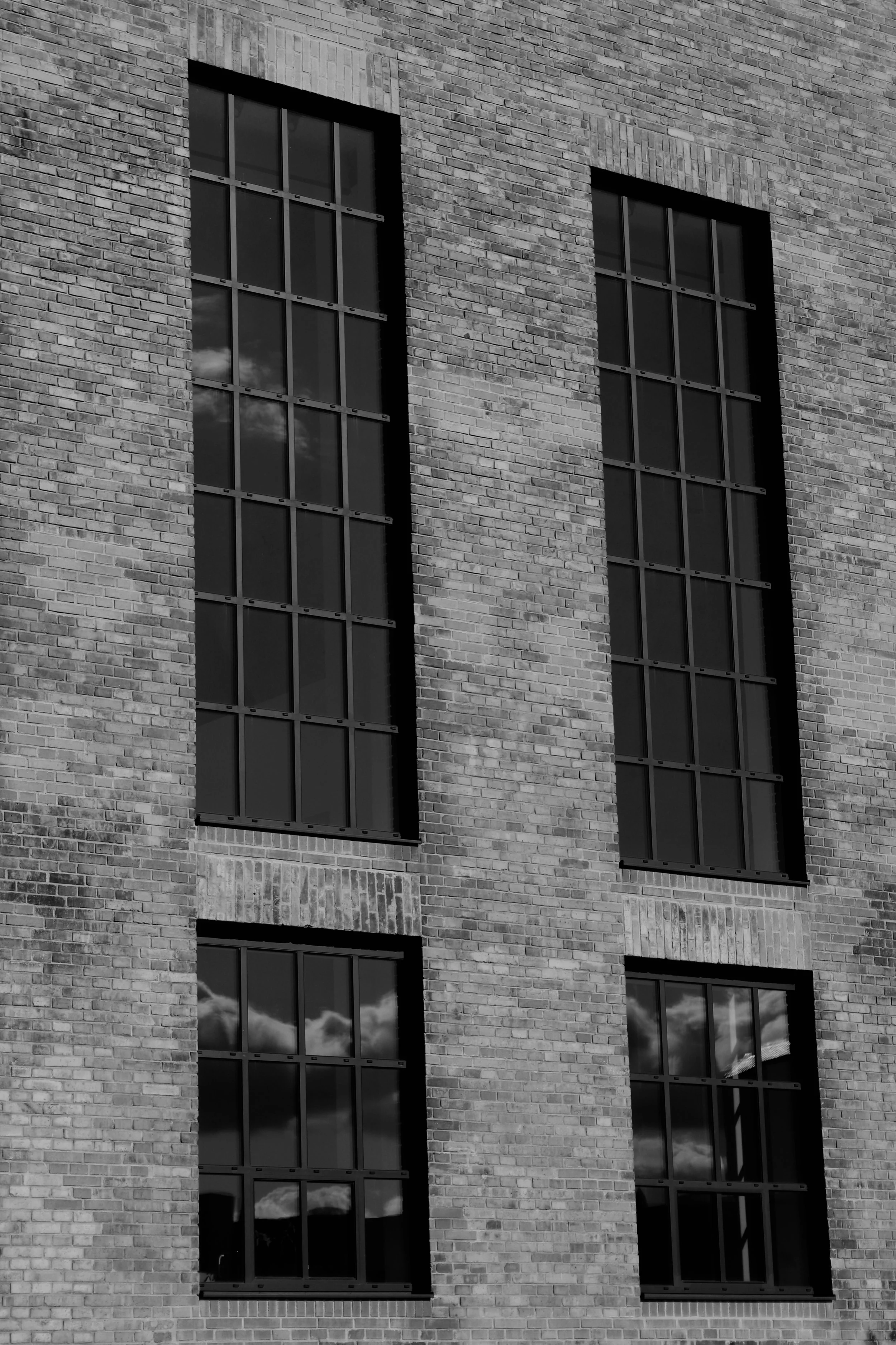 Black and white photograph of a brick building exterior with four large rectangular windows reflecting clouds and sky.