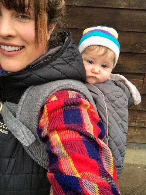 A woman smiling with a young child in a backpack carrier on her back. The child is wearing a colorful striped beanie. They are outdoors with a wooden fence in the background.