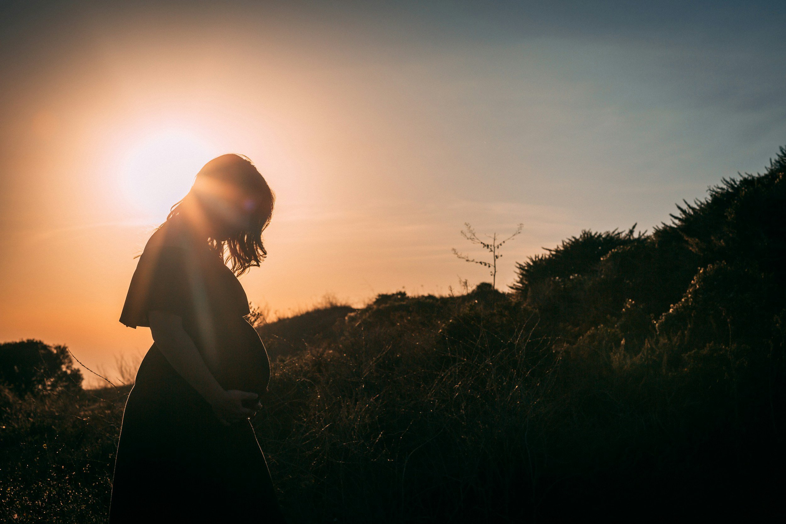 Silhouette of a pregnant woman standing outdoors during sunset, with the sun behind her, casting a glow around her.